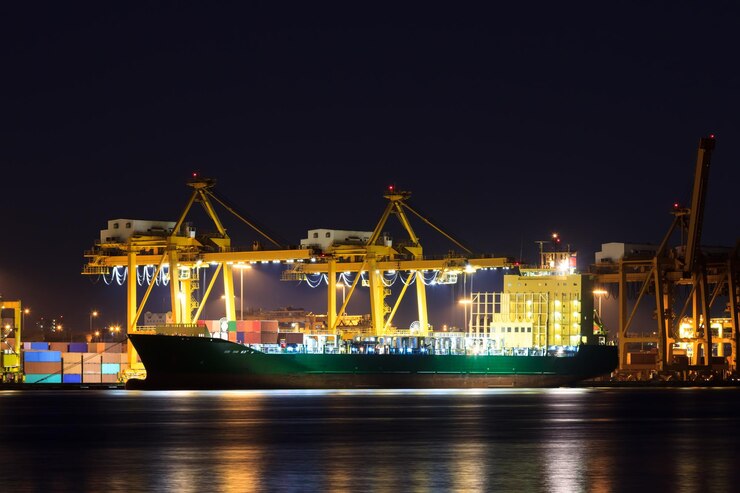 A massive cargo vessel docked in a port at night, surrounded by cranes and other industrial equipment. The ship is likely carrying Temporary Construction Space Containers, which offer numerous benefits for construction projects.