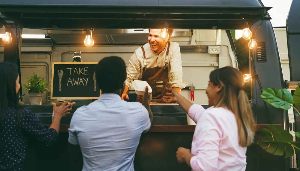 A crowd gathers in front of a food truck, surrounded by shipping containers for food services.